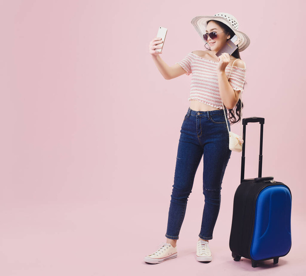 Asian pretty young girl smiling wear sunglasses and take a selfie by smartphone, Young women backpacker take a selfie and holding passport in studio pink background. Pastel pink tone filters. - Photo, Image