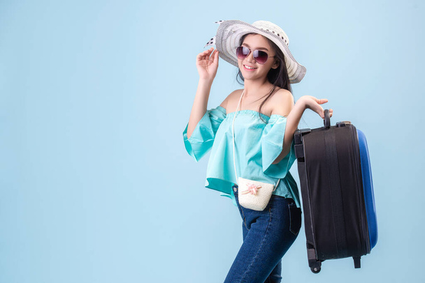 帽子をかぶって眼鏡をかけて、明るい顔をした若いアジアの女の子。スタジオブルーパステルの背景で夏に旅行する手荷物を保持。パステルブルートーンフィルター. - 写真・画像