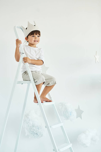 portrait of a cute little boy in crown on white ladder among clouds on a white background - Foto, imagen