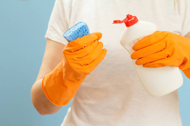 Vrouw in een t-shirt met een spons en een plastic fles wasvloeistof op de blauwe achtergrond. Was- en reinigingsconcept. - Foto, afbeelding