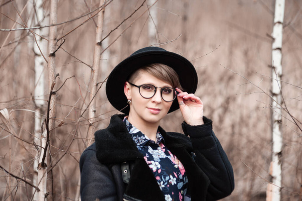Beautiful happy blonde woman in ablack hat, glasses and a jacket in autumn posing near the trees - Photo, Image