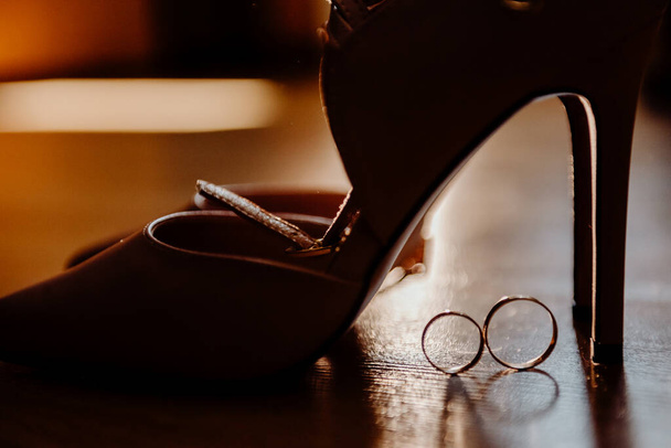 Anéis de ouro ficam ao lado do chão sob os sapatos de noivas iluminados por luz artística com destaques e reflexão de luz nas faces dos anéis
 - Foto, Imagem