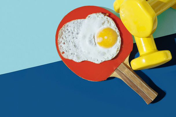 On the blue table are yellow dumbbells, a red ping pong racket, and eggs. Eggs with a thin yolk are lying on rocket. - Foto, Imagem