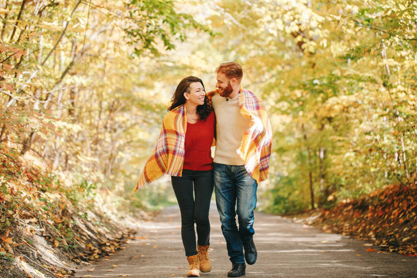Beautiful couple man woman in love. Smiling laughing boyfriend and girlfriend wrapped in yellow blanket walking in park on autumn fall day. Togetherness and happiness. Authentic real people. - Фото, зображення