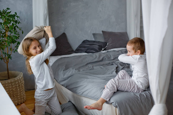 Неслухняні діти маленький хлопчик і дівчинка в піжамі билися подушкою на ліжку в сірій спальні. Їм подобається ця гра. Будинок веселий
. - Фото, зображення