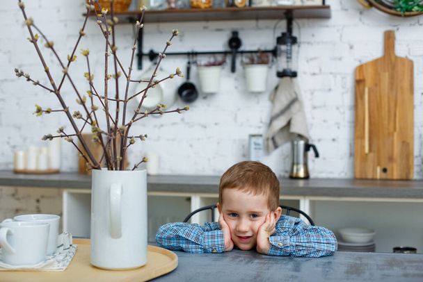 A brooding little blond boy sits at the kitchen table and looks at the camera. Cute little boy gets bored during quarantine. Sad child sitting on a chair, loneliness - Foto, Imagen