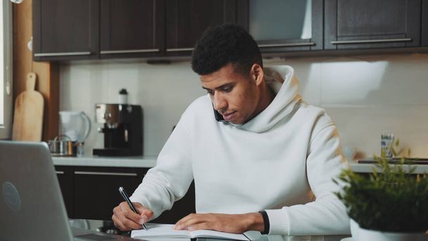 Mixed-race man speaking by smartphone and working on computer at home kitchen - Photo, image