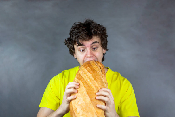 Ευτυχισμένο όμορφο νεαρό αγόρι κρατά και τρώει φρεσκοψημένο ψωμί. Πεινασμένο αγόρι δαγκώνει μεγάλο καρβέλι, σε γκρι φόντο. - Φωτογραφία, εικόνα