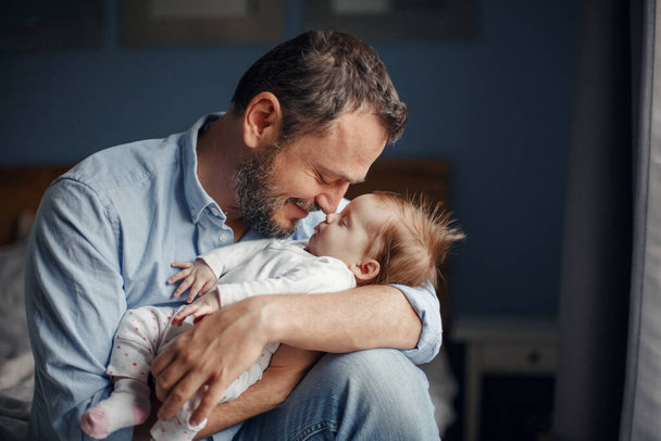 中年白人の父親が新生児の女の子にキスをする。親はロッキングチャイルドの娘の息子を手に持ってる。本物のライフスタイル子育ての父親の瞬間。シングルファーザー家庭生活. - 写真・画像