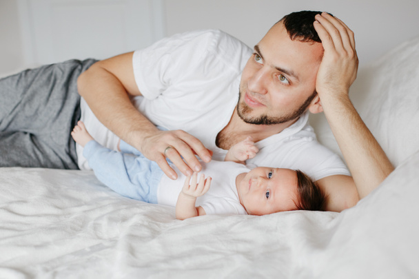 幸せな白人の父親は、自宅のベッドルームで新生児と横たわっています。男の親は子供の娘の息子を保持。本物のライフスタイルの率直な瞬間。誇り高い若いお父さん。父の日. - 写真・画像