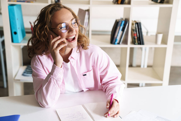 Photo of excited blonde woman wearing eyeglasses talking on cellphone while doing homework and sitting in room - Fotoğraf, Görsel