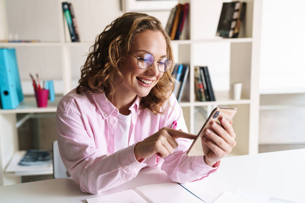 Photo of cheerful nice woman wearing eyeglasses using cellphone while doing homework and sitting in room - Foto, Imagen