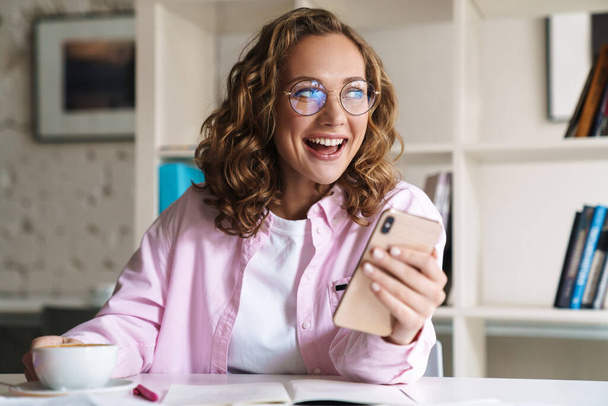 Photo of smiling nice woman wearing eyeglasses using cellphone and drinking coffee while doing homework in room - Fotoğraf, Görsel