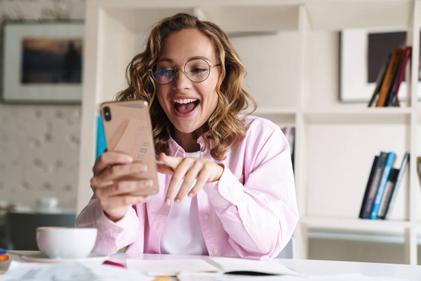 Photo of smiling nice woman wearing eyeglasses making video call on cellphone while doing homework in room - Foto, Imagen