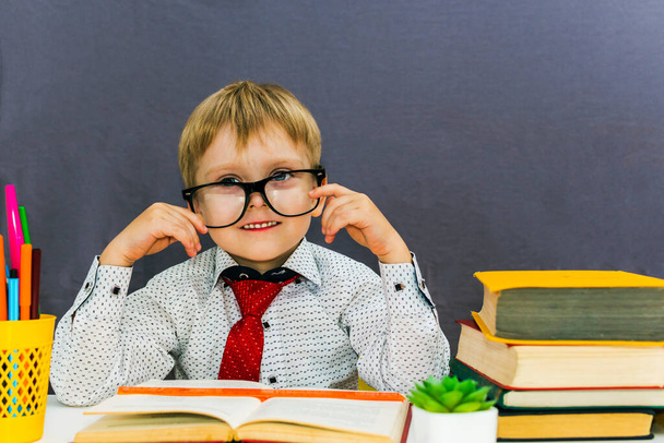 slimme grappige jongen met bril aan een tafel met boeken op een grijze achtergrond - Foto, afbeelding