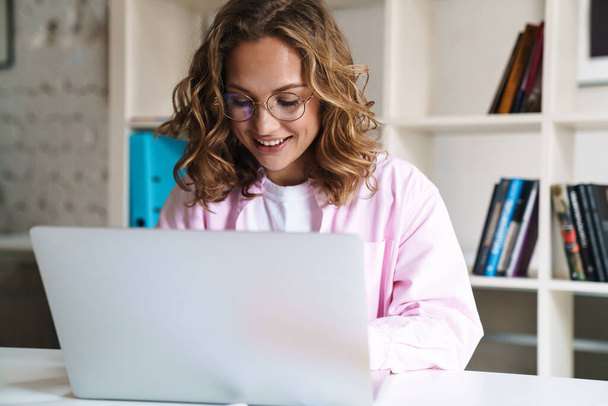 Photo of joyful blonde woman wearing eyeglasses smiling and using laptop while sitting in cafe - Fotoğraf, Görsel