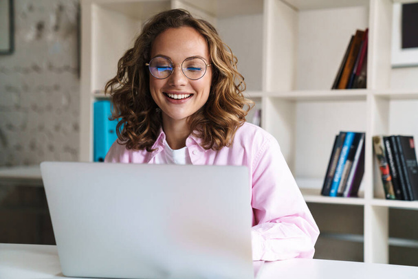 Photo of joyful blonde woman wearing eyeglasses smiling and using laptop while sitting in cafe - 写真・画像