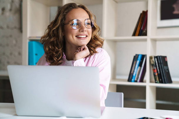 Photo of joyful blonde woman wearing eyeglasses smiling and using laptop while sitting in cafe - Foto, Imagen