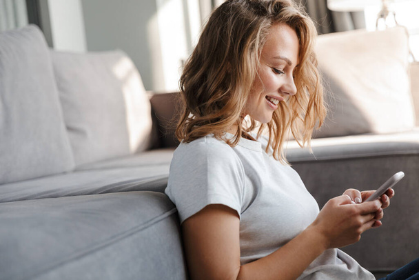 Image of happy caucasian woman in basic t-shirt smiling and using cellphone while sitting on floor at living room - Photo, Image