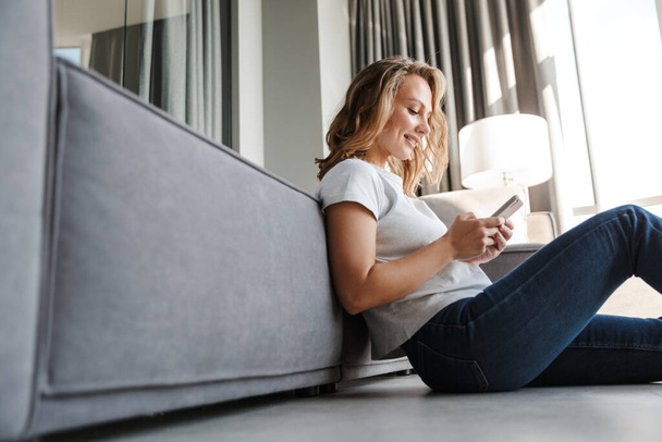 Image of happy caucasian woman in basic t-shirt smiling and using cellphone while sitting on floor at living room - Fotó, kép