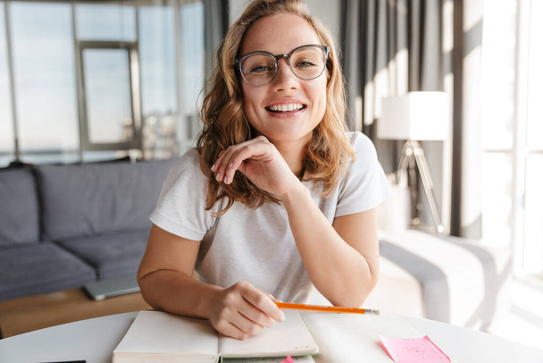 Image of happy caucasian woman in eyeglasses smiling and making notes in diary while sitting at table - Foto, Bild