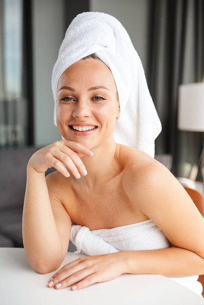 Photo of young joyful woman wrapped in white towels smiling at camera while sitting in room after shower - Foto, immagini