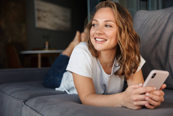 Smiling pretty blonde young woman wearing casual clothes using mobile phone while relaxing on a couch at home - Foto, Imagen