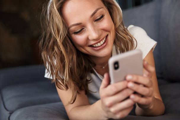 Image of smiling blonde woman using cellphone and wireless headphones while lying on sofa at living room - 写真・画像