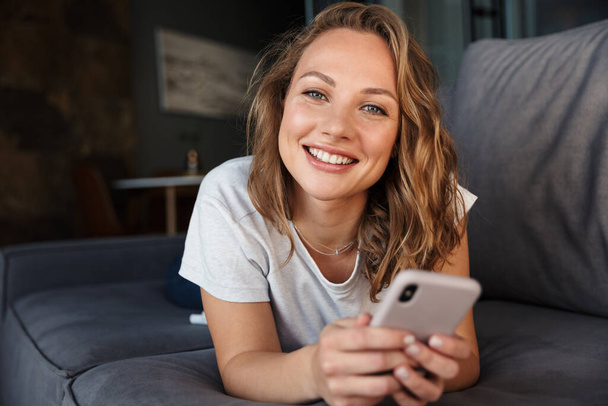 Image of happy blonde woman in basic t-shirt smiling and using cellphone while lying on sofa at living room - Φωτογραφία, εικόνα
