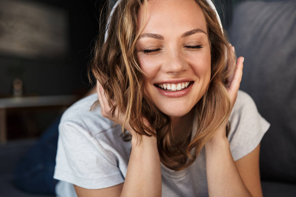 Image of smiling blonde woman using wireless headphones while lying on sofa at living room - Zdjęcie, obraz