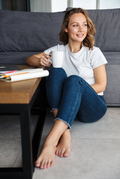 Image of happy attractive woman in basic t-shirt smiling and drinking coffee while sitting on floor at living room - Fotó, kép