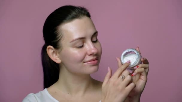 Sexy brunette caucasian woman in neutral clothes standing on a pink background. She apply cream on her clean face (no mascara). Cosmetic concept - Footage, Video