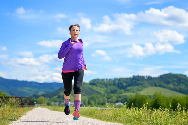 Mulher correndo ao longo de uma estrada rural em sol de primavera durante seu treino diário em uma visão de baixo ângulo para conceitos de fitness e estilo de vida ativo
 - Foto, Imagem