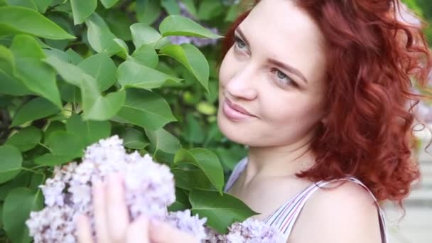 Mujer caucásica linda pelirroja en vestido de verano relajante en el parque, oliendo flores lila y sonriendo felizmente. Retrato de la cabeza en un soleado día de primavera
 - Metraje, vídeo