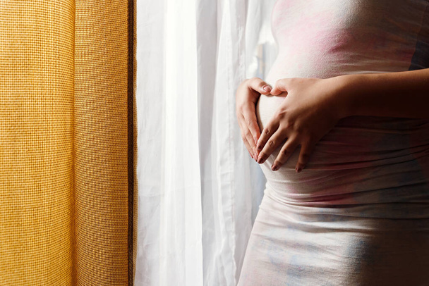 беременная молодая женщина, перед окном с большим освещением, комфортно одета, держит большой живот, беременная ожиданиями, День матери, руки на животе с сердечной фигурой
. - Фото, изображение