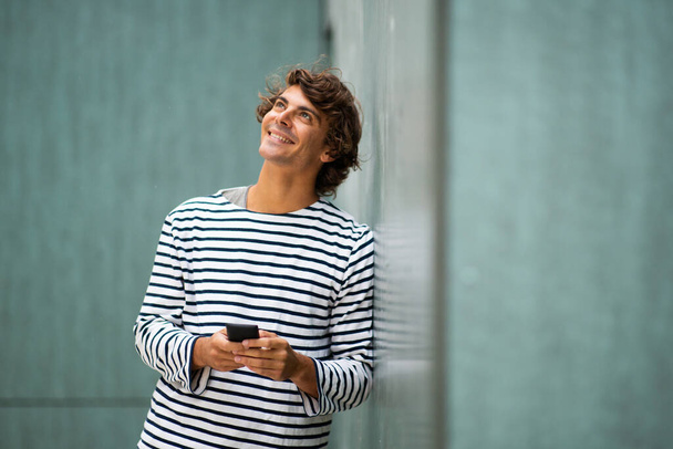 Portret uśmiechnięty młody człowiek opierający się o zieloną ścianę trzymając telefon komórkowy - Zdjęcie, obraz