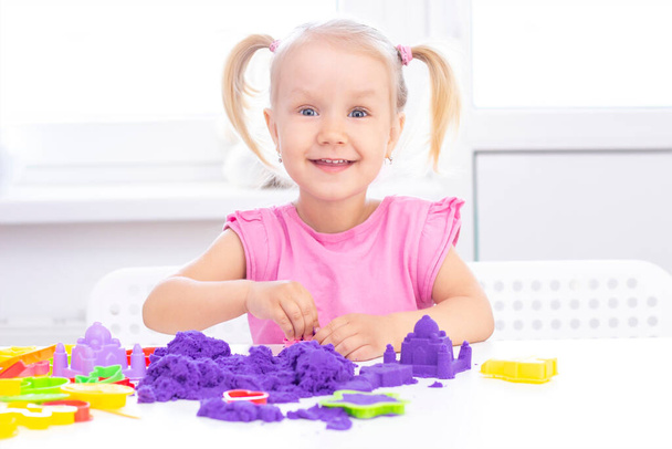 happy Girl plays kinetic sand in quarantine. Blond beautiful girl smiles and plays with purple sand on a white table. - Photo, Image