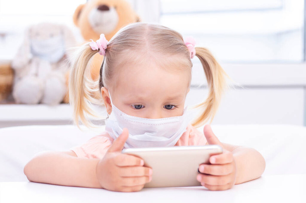girl in a protective medical mask using a mobile phone, a smartphone for video calls, talks with relatives, a girl sits at home, online computer webcam, making a video call. - Φωτογραφία, εικόνα