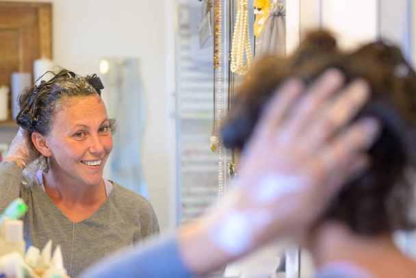 Frau färbt ihr Haar mit Haarfärbung zu Hause lächelnd sich selbst im Badezimmerspiegel mit Fokus auf die Reflexion - Foto, Bild