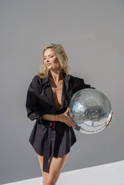 Young blond model in black dress and high heels posing with disco-ball on grey studio background   - Foto, Imagen