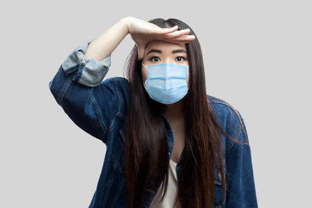 Portrait of attentive beautiful brunette asian young woman with surgical medical mask in blue denim jacket standing with hand on forehead and looking. indoor studio shot, isolated on grey background. - 写真・画像