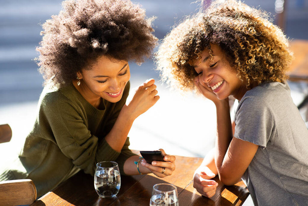 Портрет двох молодих афроамериканських жінок, які сидять у кафе на вулиці і дивляться на мобільний телефон. - Фото, зображення