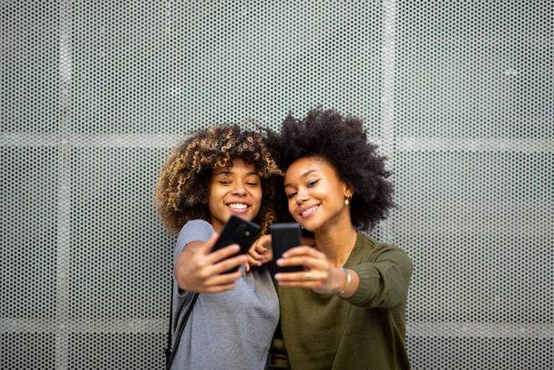 Πορτρέτο δύο χαρούμενες νεαρές μαύρες γυναίκες που παίρνουν selfie με κινητά τηλέφωνα  - Φωτογραφία, εικόνα