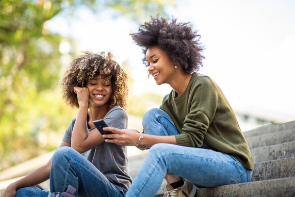 Seitenporträt zwei junge afrikanisch-amerikanische Frauen, die auf den Stufen vor dem Haus sitzen und auf ihr Handy schauen - Foto, Bild