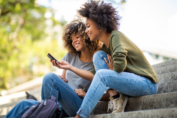 Retrato lateral de dos jóvenes afroamericanas sentadas en escalones afuera mirando el teléfono celular
 - Foto, imagen