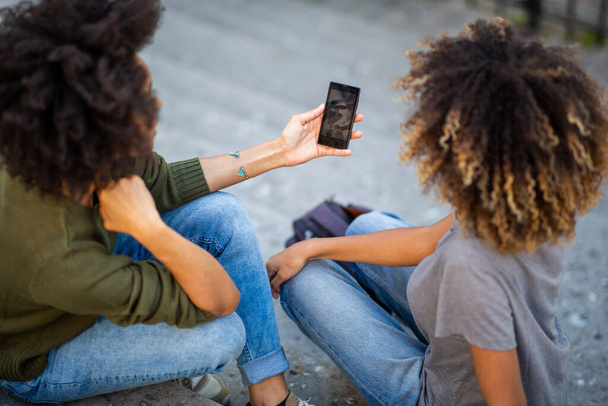 Portrait de derrière de deux jeunes femmes afro-américaines assises sur des marches à l'extérieur regardant le téléphone portable
 - Photo, image