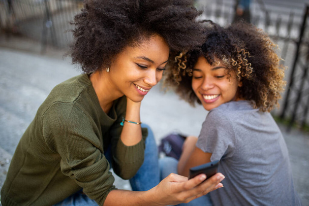 Portrait deux jeunes femmes noires heureuses assises sur des marches à l'extérieur regardant le téléphone mobile
 - Photo, image