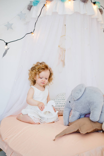 Ένα κοριτσάκι παίζει σε μια λευκή ηλιόλουστη κρεβατοκάμαρα. Ένα παιδί με παιχνίδια. Παιχνίδι ρόλων για μικρά παιδιά. Ένα παιδί με ένα βελούδινο παιχνίδι ελέφαντα. Παιδικό δωμάτιο εσωτερικό - Φωτογραφία, εικόνα