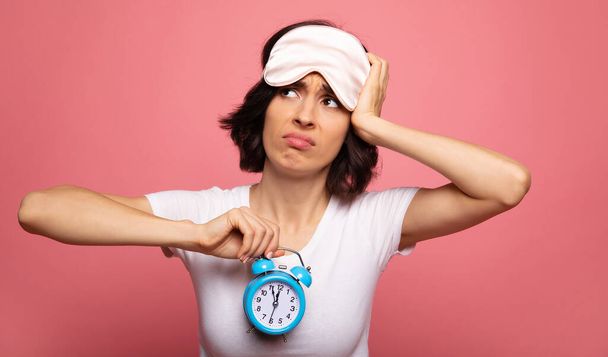 Troubles with sleep. Close-up photo of dissatisfied woman in a face mask, who is touching her head and looking up, while holding a clock in her right arm. - Foto, immagini