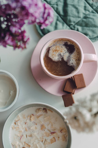 Beautiful pink cup with coffee and milk chocolate on warm grey plaid. Tasty breakfast in bed with lilac flowers in spring morning - Photo, Image